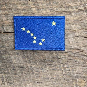 以下が含まれることがあります： 青い刺繍のパッチで、北斗七星の黄色のアウトラインが描かれています。パッチは木製の背景の上にあります。