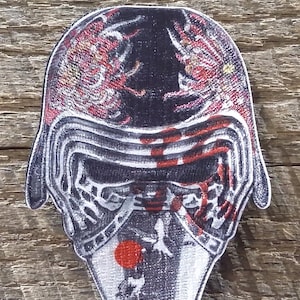 May include: An embroidered patch of a Star Wars character's helmet in black and white with red and orange accents. The helmet features a red sun and a white bird design on the front.
