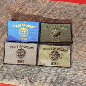 Puede incluir: Cuatro parches bordados con el sello del estado de Oregón. Los parches son de diferentes colores y patrones, incluyendo azul, marrón y camuflaje.