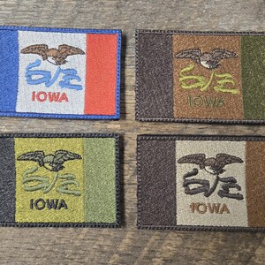 Puede incluir: Cuatro parches bordados con el diseño de la bandera del estado de Iowa. Cada parche tiene forma rectangular con borde. Los diseños incluyen una combinación de colores azul, blanco y rojo, junto con el nombre del estado y un emblema de ave.
