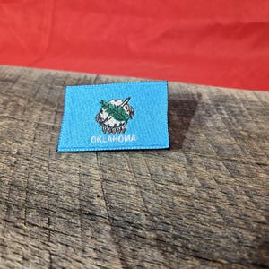 May include: A rectangular patch featuring the Oklahoma state flag. The patch has a light blue background with the state seal in the center. The word "OKLAHOMA" is embroidered in white at the bottom. The patch is on a wooden surface.