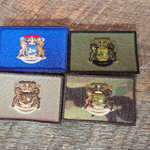May include: Four rectangular embroidered patches with a coat of arms design. The patches are in blue, olive green, tan, and camouflage patterns. The coat of arms features a shield, two figures, and a crown.