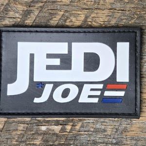 May include: A rectangular black patch with the words "JEDI JOE" in white block letters. The "JEDI" is stacked above "JOE". A small blue star is next to the "J" in "JEDI". Below "JOE" are three horizontal stripes in red, white, and blue.