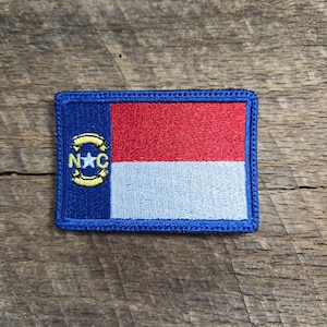 May include: A blue embroidered patch featuring the North Carolina state flag. The flag has a red and white stripe with a blue star and yellow letters "NC" in a circle.