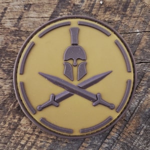 May include: A circular patch featuring a raised dark brown Spartan helmet centered above two crossed swords. The design is set against a tan background with a dark brown circular border, creating a distinct contrast between the elements.