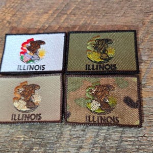 Op de afbeelding: Vier rechthoekige patches met het zegel van de staat Illinois. De patches hebben verschillende achtergrondkleuren: wit, olijfgroen, beige en camouflage. Elke patch heeft het woord "ILLINOIS" in een donkere kleur geborduurd.
