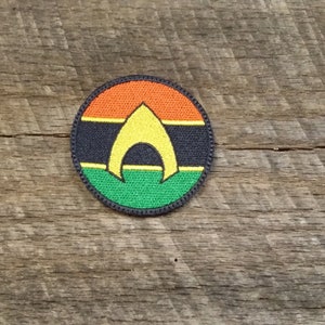 May include: A round, black, green, yellow, and orange embroidered patch with the Aquaman logo. The logo is a yellow trident with a black outline.
