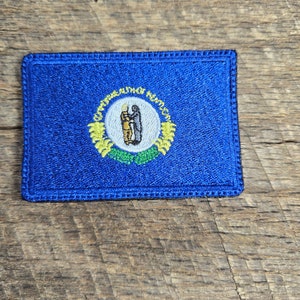 May include: A blue embroidered patch with the state seal of Kentucky. The seal features a  yellow wreath with the words "Commonwealth of Kentucky" and two figures shaking hands.
