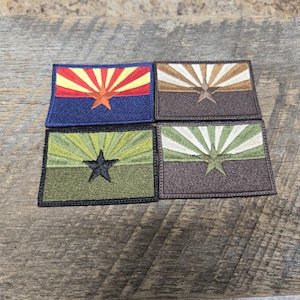 Puede incluir: Cuatro parches bordados de la bandera del estado de Arizona en diferentes combinaciones de colores. Los parches presentan un diseño de rayos de sol rojo, amarillo y blanco con una estrella en el centro. Los parches están sobre una superficie de madera.