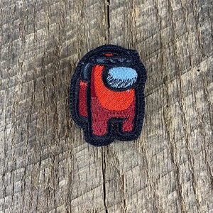 May include: A red and black embroidered patch of an Among Us character. The character is wearing a black visor and has a black outline.