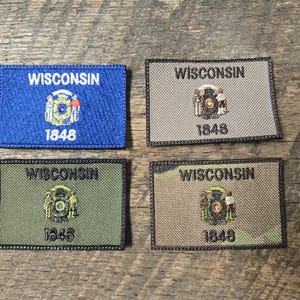 Puede incluir: Cuatro parches rectangulares de la bandera del estado de Wisconsin en azul, gris, verde y camuflaje. Cada parche presenta la palabra "WISCONSIN", el sello estatal y el año "1848". Los parches están sobre una superficie de madera.