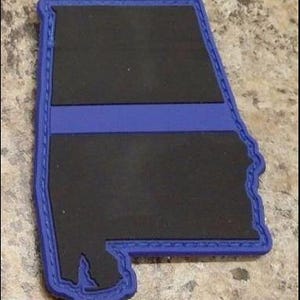 May include: A black and blue patch in the shape of the state of Alabama. The patch has a blue border and a blue stripe across the middle. The patch is a representation of the Thin Blue Line.