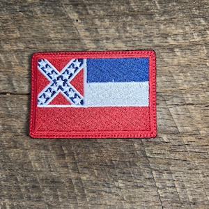 Pode incluir: Um remendo bordado com a bandeira confederada do estado do Mississippi. O remendo é vermelho, branco e azul e está sobre um fundo de madeira.