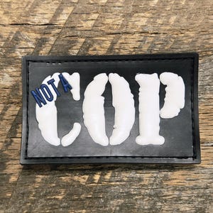 Op de afbeelding: Een zwarte rechthoekige patch met de woorden "NOT A COP" in witte, gestileerde letters. Het woord "NOT A" is blauw. De patch is geplaatst tegen een houten achtergrond.