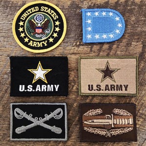 May include: A collection of six embroidered patches including a circular United States Army emblem, a blue D-shaped patch with white stars, two rectangular U.S. Army patches in black and tan, a crossed swords insignia, and a brown bayonet patch.