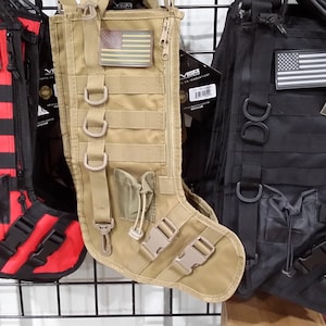May include: Three tactical Christmas stockings in red, beige, and black, each with an American flag patch. The stockings feature multiple straps, buckles, and pockets, and are designed to resemble military gear.