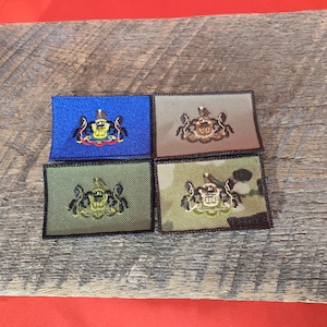 Könnte beinhalten: Vier gestickte Aufnäher mit dem Staatssiegel von Pennsylvania. Die Aufnäher sind in verschiedenen Farben und Stoffen gehalten, darunter Blau, Braun, Grün und Tarnmuster.