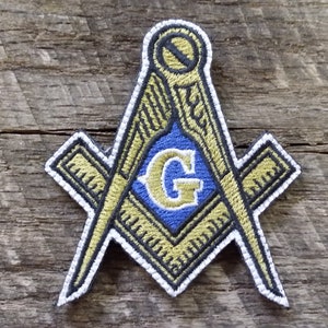 May include: A gold and blue embroidered patch featuring a square and compass with a 'G' in the center. The patch is on a wooden background.