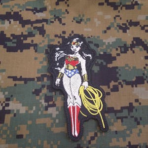 May include: Embroidered patch featuring Wonder Woman in her iconic red, blue, and gold costume. She is holding her lasso of truth in her right hand. The patch is set against a camouflage background.