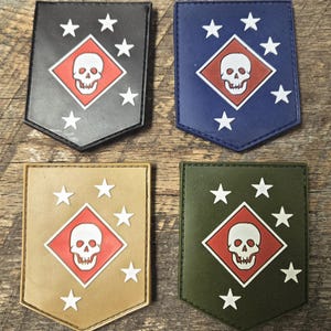 May include: Four shield-shaped morale patches in black, blue, tan, and olive green. Each patch features a central red diamond containing a white skull, surrounded by five white stars arranged in an arc. The patches have a flat, smooth finish.