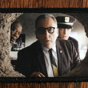 May include: A black and white embroidered patch featuring a scene from a movie. Three men are shown, one with glasses and a suit, another in a police uniform, and a third man partially obscured by a wall. The scene appears to be from a tense moment in the film.