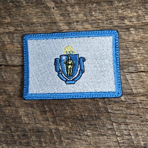 May include: A blue and white embroidered patch featuring the state seal of Massachusetts. The seal depicts a Native American holding a bow and arrow, surrounded by a blue and gold shield.