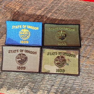 Könnte beinhalten: Vier gestickte Aufnäher mit dem Siegel des Bundesstaates Oregon, dem Text "STATE OF OREGON" und "1859". Die Aufnäher sind in Blau, Olivgrün, Beige und Camouflage-Mustern auf einer Holzoberfläche dargestellt.