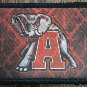 Peut inclure: Écusson rectangulaire à bordure noire avec un motif rouge et blanc. Le motif comprend un éléphant stylisé et la lettre "A" dans un style universitaire audacieux. Le fond a un aspect texturé et à motifs.