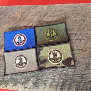 Könnte beinhalten: Vier gestickte Aufnäher mit dem Staatssiegel von Virginia. Die Aufnäher sind in verschiedenen Farben und Mustern gehalten, darunter Blau, Grün, Braun und Tarnmuster.