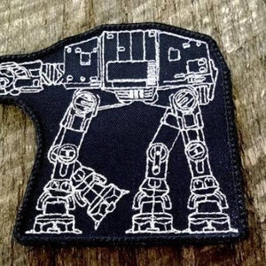 May include: Black patch featuring a white outline of an AT-AT walker from Star Wars. The patch has a black background with white stitching around the edges. The design is a detailed illustration of the vehicle.