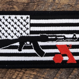 May include: A rectangular embroidered patch featuring a black and white American flag design. A black silhouette of an assault rifle is centred across the flag, and a red geometric logo is positioned in the lower right corner of the patch.