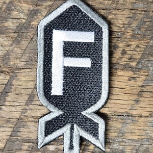 May include: Black and gray embroidered patch with a white border and a white "F" in the center. The patch has a unique shape with a pointed top and a stylized bottom. The patch is on a wooden surface.