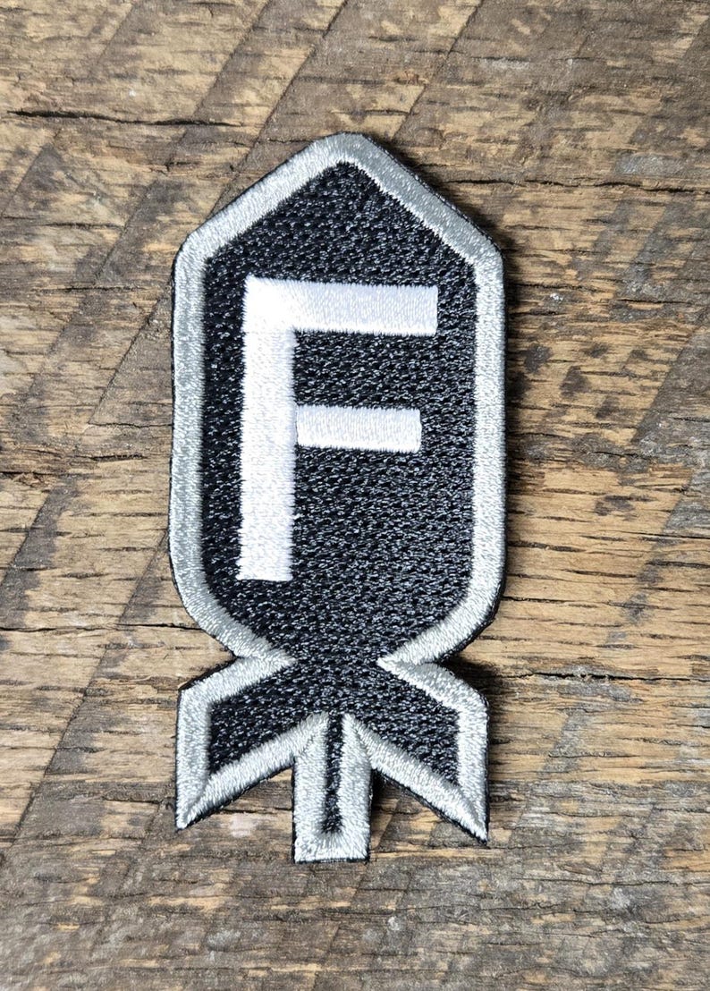 May include: Black and gray embroidered patch with a white border and a white "F" in the center. The patch has a unique shape with a pointed top and a stylized bottom. The patch is on a wooden surface.