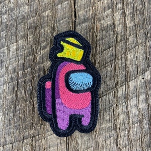 Puede incluir: Parche bordado con un personaje del juego Among Us. El personaje tiene un cuerpo rosa, piernas moradas y una visera azul. Una corona amarilla está en la cabeza, todo con un contorno negro.