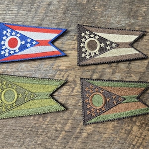 May include: Four embroidered patches in the shape of the Ohio state flag. One patch is blue, red, and white. The other three patches are in shades of brown and green. The patches are on a wooden surface.
