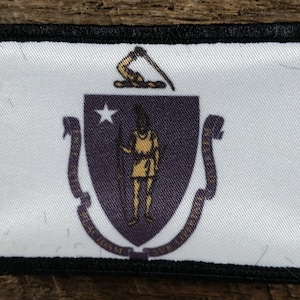 May include: A white patch with a black border featuring the state seal of Massachusetts. The seal depicts a Native American man holding a bow and arrow, with a star above him. The seal is surrounded by a purple shield and two gold scrolls with the words "Ense petit placidam sub libertate quietem" on them.