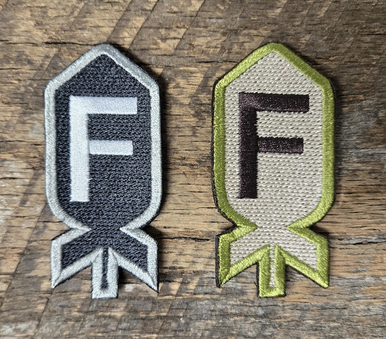 May include: Two embroidered patches with a shield-like shape and a stylized letter "F". One patch is black with a white "F" and silver trim. The other is beige with a brown "F" and green trim. Both have a bomb-like base.