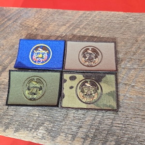Puede incluir: Cuatro parches rectangulares con emblemas circulares bordados. Uno es azul, los otros son beige, verde oliva y camuflaje. Los emblemas presentan un diseño central dentro de un borde circular. Los parches están sobre una superficie de madera envejecida.