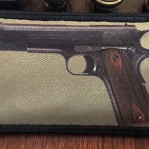 May include: A rectangular patch featuring a detailed image of a handgun with a brown grip. The patch has a black border and a tan background. The handgun is depicted in a side profile, showcasing its design.