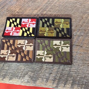May include: Four rectangular patches with the Maryland state flag design. Each patch features a different color scheme, including black and yellow, green and brown, and various shades of brown and green. The patches are displayed on a wooden surface.
