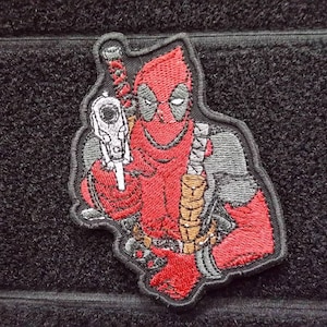 以下が含まれることがあります： 銃を持ったデッドプールが刺繍されたパッチ。赤と黒の配色で、生地製で細部までこだわったデザインです。