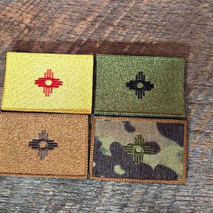 Könnte beinhalten: Vier rechteckige Aufnäher mit dem Zia-Symbol von New Mexico. Die Aufnäher sind in Gold mit einem roten Symbol, Olivgrün mit einem schwarzen Symbol, Braun mit einem schwarzen Symbol und Tarnfarben mit einem grünen Symbol. Auf einer Holzoberfläche.