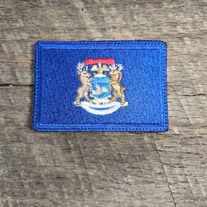 Pode incluir: Um remendo bordado azul com o selo do estado de Michigan. O selo apresenta um escudo com uma fita vermelha, dois alces e uma águia branca.