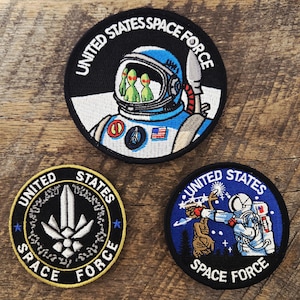 May include: Three circular embroidered patches featuring United States Space Force designs. One shows an astronaut with green aliens in the helmet, one displays a central silver emblem, and one depicts an astronaut reaching toward a creature.