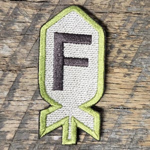 May include: Embroidered patch featuring the letter "F" in dark brown, centered on a light gray background. The patch has a lime green border and a stylized shape resembling a bomb. The patch is set against a weathered wood background.