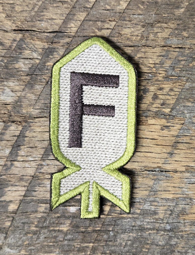 May include: Embroidered patch featuring the letter "F" in dark brown, centered on a light gray background. The patch has a lime green border and a stylized shape resembling a bomb. The patch is set against a weathered wood background.