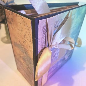 Puede incluir: Un álbum de recortes hecho a mano con una cubierta texturizada y estampada en tonos marrón, azul y beige. Los bordes son negros y una cinta color crema está atada alrededor del lomo. Una cinta de color crema sobresale de la parte superior.