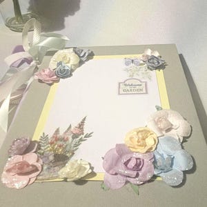 Puede incluir: Un álbum de recortes decorativo con adornos florales. La cubierta es gris, con un interior blanco y amarillo. El álbum está adornado con varias flores de papel de colores pastel, mariposas y un letrero que dice "Welcome to my Garden."
