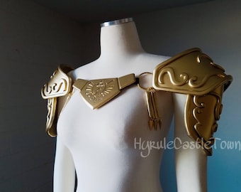 May include: Gold-colored shoulder armor with a decorative design. The armor is worn on a white mannequin. The armor has a large, triangular piece in the center with a stylized bird design.