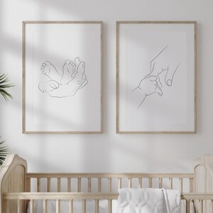 Puede incluir: Dos impresiones art&iacute;sticas enmarcadas con dibujos lineales. Una representa una mano acunando pies de beb&eacute;, la otra una mano sosteniendo la mano de un beb&eacute;. Los marcos son de madera clara y el arte se exhibe en una pared blanca. El tema es beb&eacute; y familia.